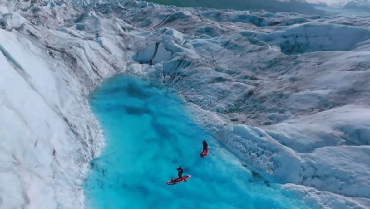 Two kayakers paddling through a turquoise glacial meltwater lake surrounded by towering white ice walls and blue crevassed glacier.