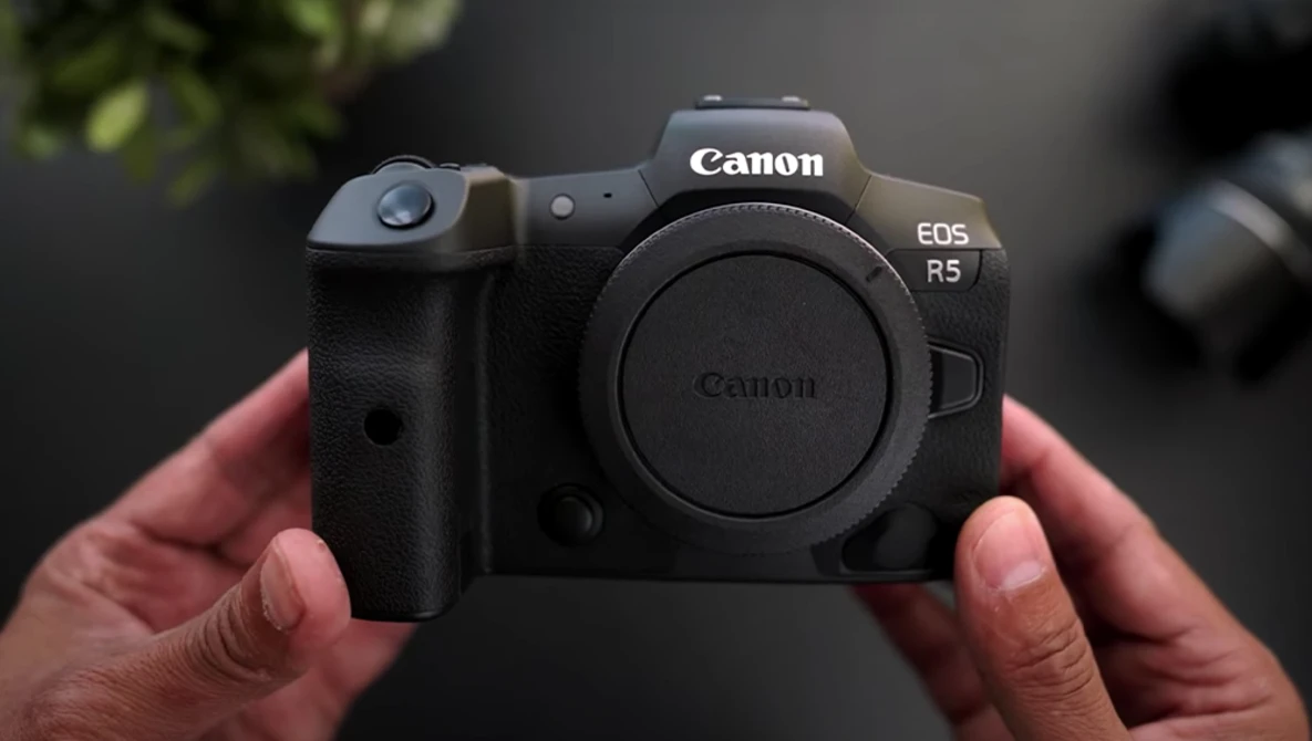 Canon EOS R5 mirrorless camera held in hands, body only, against blurred background.
