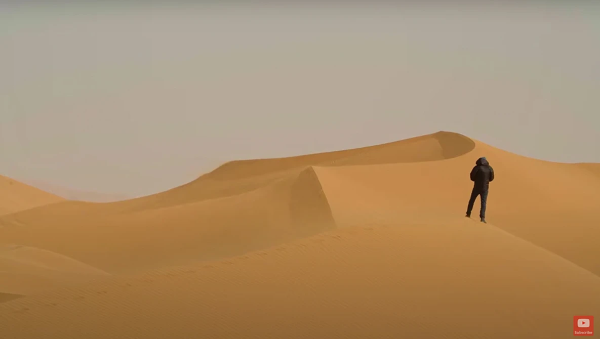 Solitary figure in dark clothing standing on a massive sand dune under hazy sky.