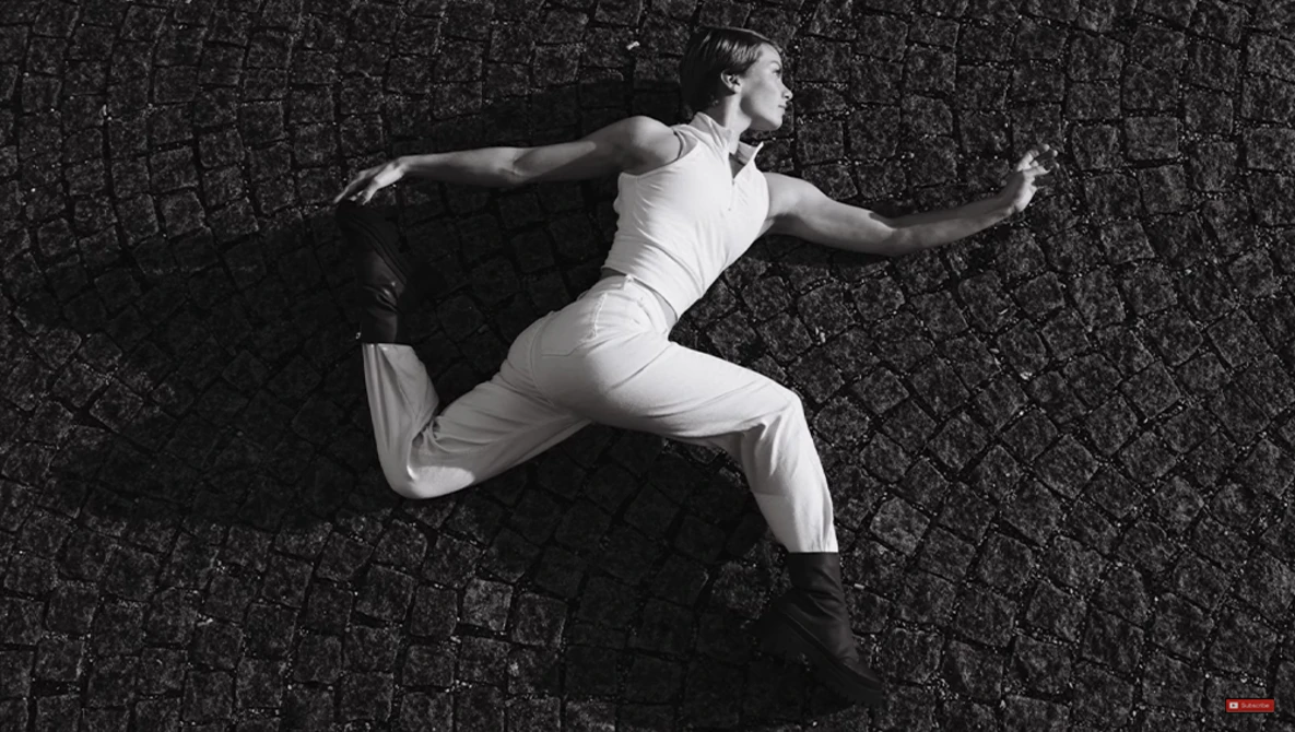 Male dancer in white outfit performing a dynamic leap against textured dark wall.