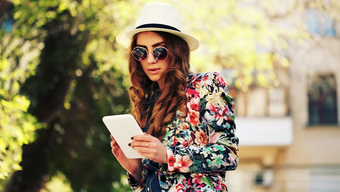 Woman in floral jacket and panama hat holding tablet in sunlit outdoor setting.