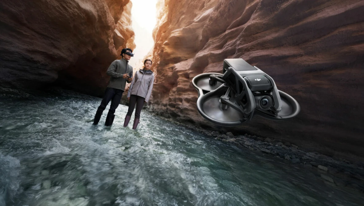 Two people standing in shallow water between towering canyon walls with a drone hovering nearby.