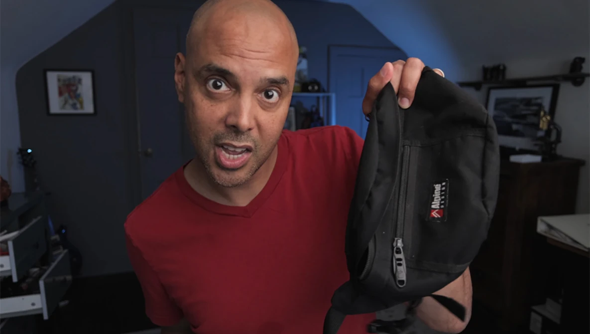Man in red shirt holding black camera bag toward camera with surprised expression.