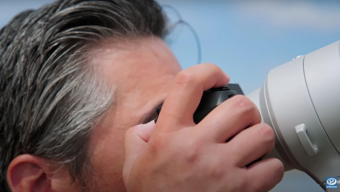 Photographer holding a telephoto lens to his eye, composing a shot against a blue sky.