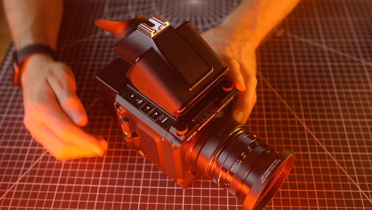 Medium format camera with attached lens held by photographer's hands against orange grid backdrop.