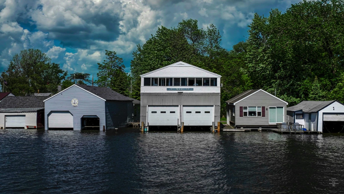 Waterfront boathouses with white garage doors reflected in calm water beneath blue sky.