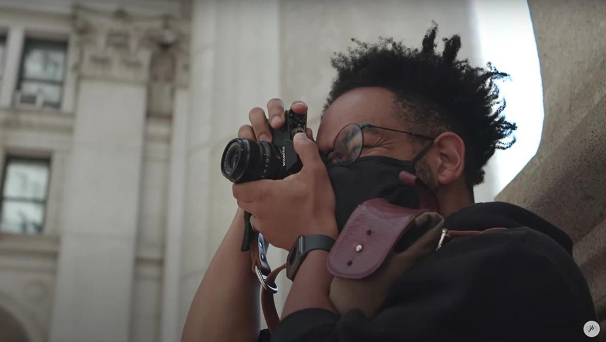 Photographer wearing glasses and face mask looking through a compact camera with urban architecture in background.