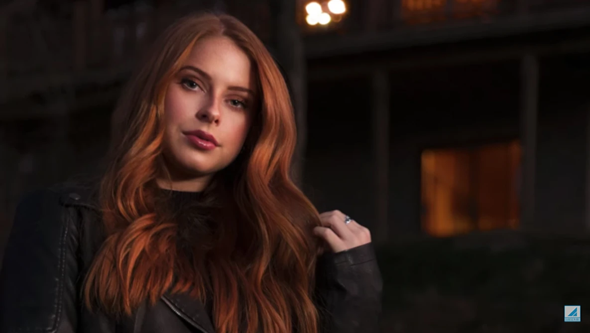 Woman with long reddish-brown hair wearing a black leather jacket, photographed outdoors at night with warm ambient lighting.