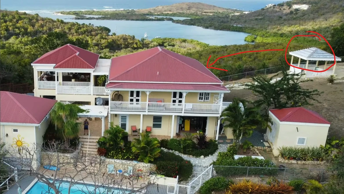 Aerial view of a large residential property with red metal roofs overlooking a coastal bay and forested hillside.