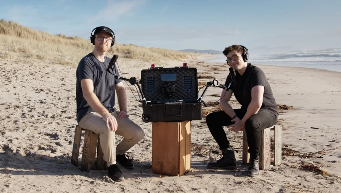Two photographers sit on wooden crates beside a black equipment case on a beach, ocean and dunes in background.