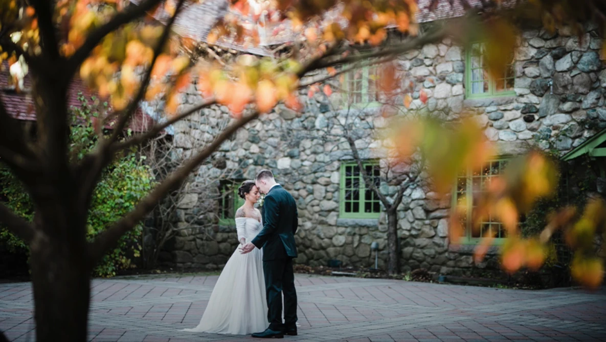 Bride and groom standing together in front of a stone building, framed by autumn foliage.