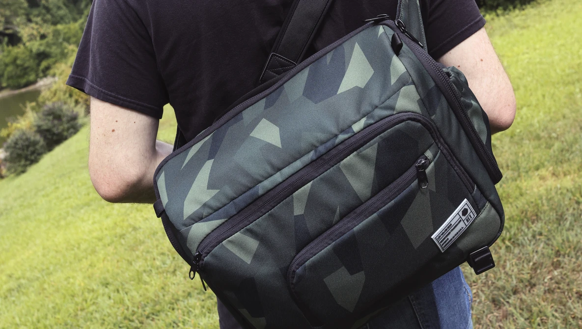 Person wearing a black and gray camo messenger bag with shoulder strap across their chest outdoors.