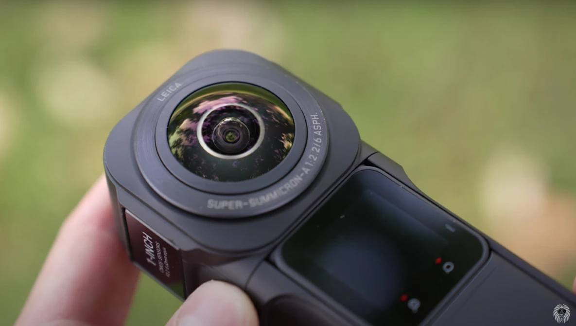 Close-up of a compact action camera with a wide-angle lens held in someone's hand against a blurred green background.