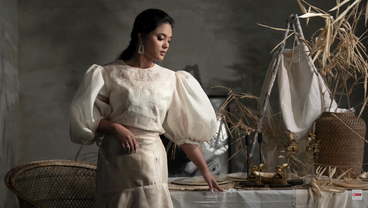 Woman in cream-colored puffed-sleeve dress posed indoors among woven baskets and dried grasses.