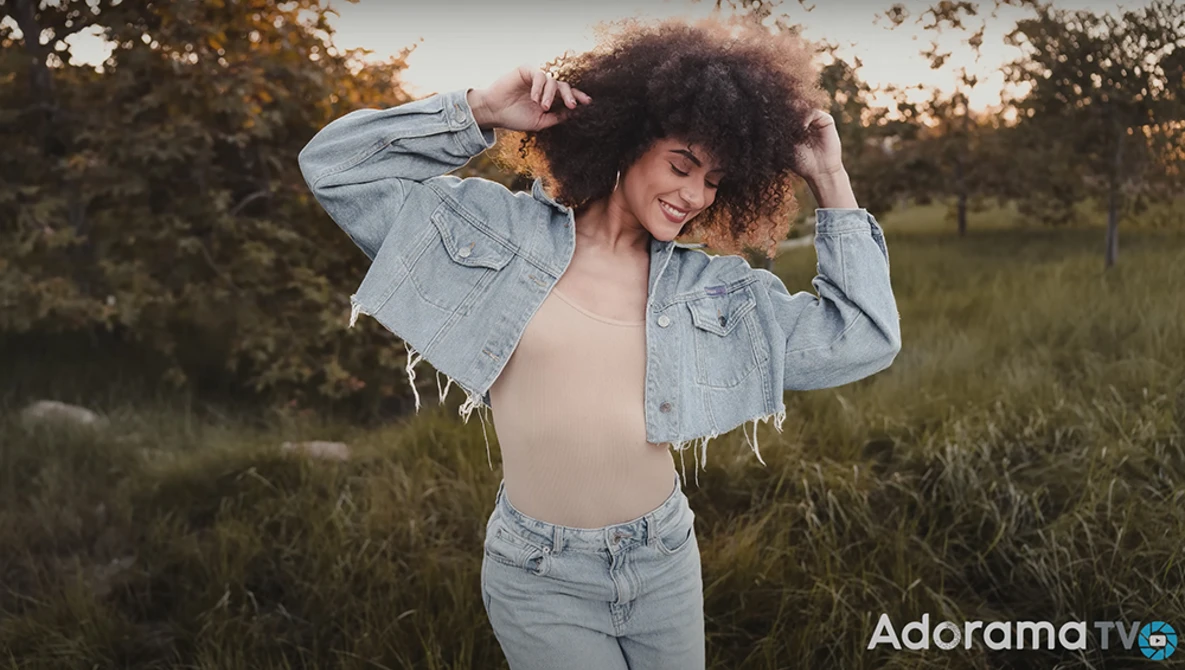 Woman with curly hair posing in an open field at golden hour, wearing an open denim jacket and jeans.