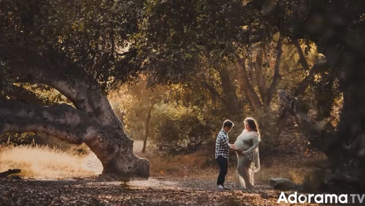 Two people standing beneath a large, gnarled tree on a sun-dappled path in a wooded area.