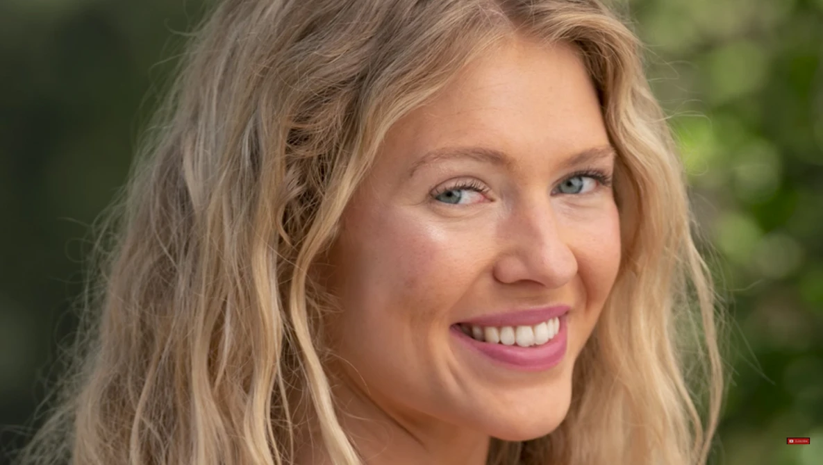 Woman with long blonde wavy hair smiling at camera outdoors with blurred green background.
