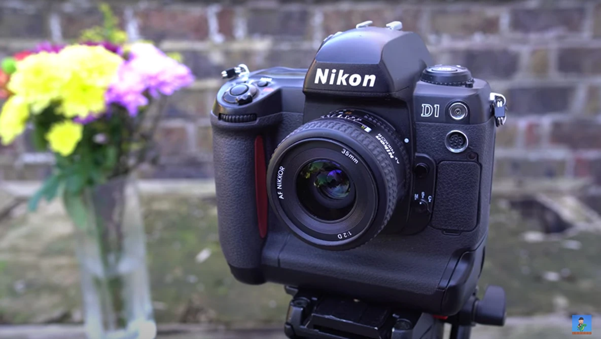 Nikon D1 DSLR camera with battery grip mounted on tripod, displayed against brick wall backdrop with flowers in vase.