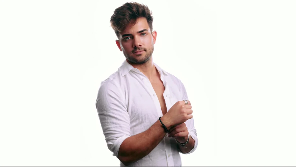 Man in light pink button-up shirt against white background, adjusting cuff with composed expression.