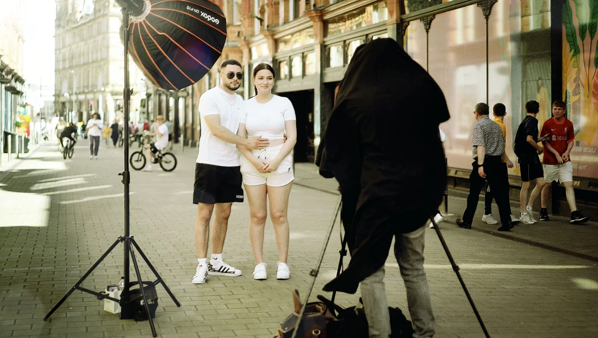 Two people posing on a city street while a photographer shoots with a large reflector and studio lighting setup.