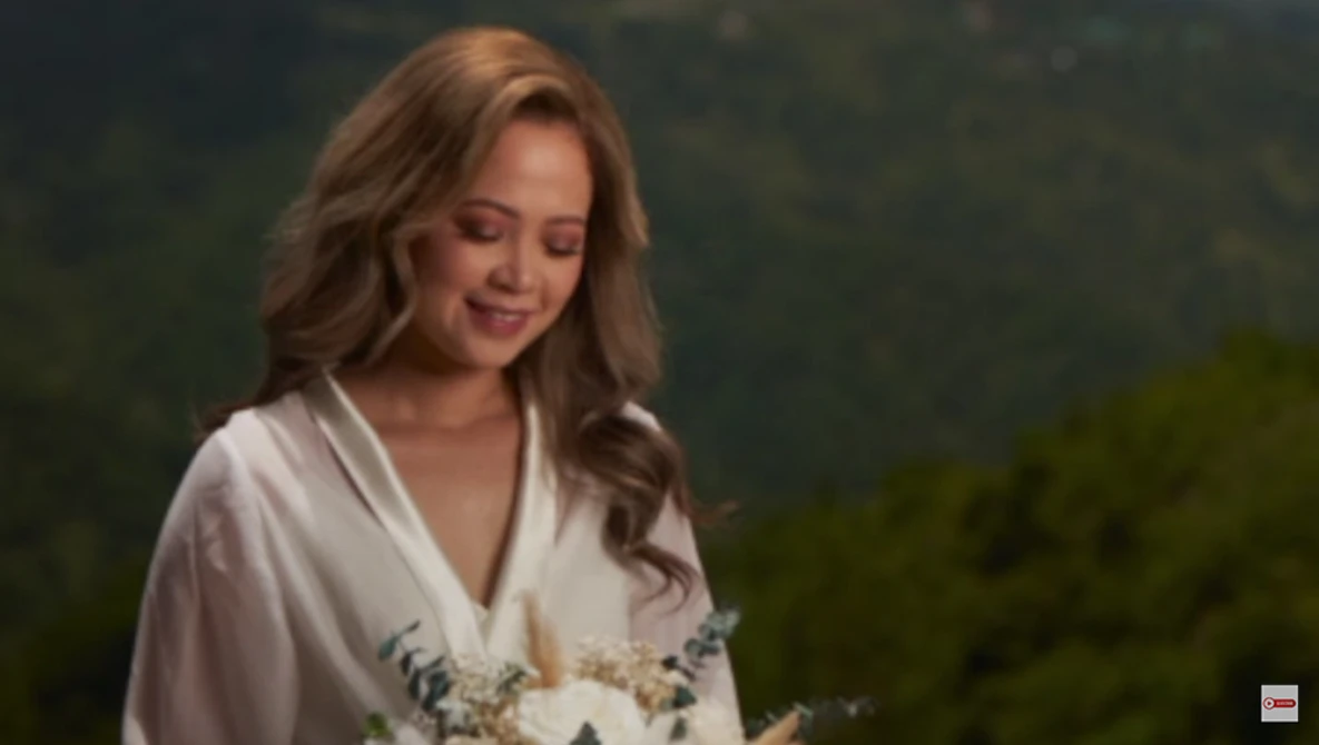 Woman in white robe holding white flowers, looking downward with soft smile outdoors.