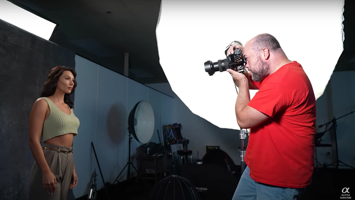 Photographer in red shirt operating cinema camera on tripod while shooting female model in studio with professional lighting setup.