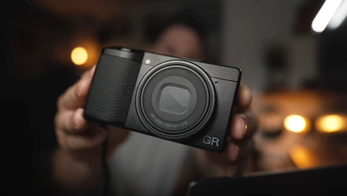 Hands holding a black compact digital camera with prominent lens and GR marking against blurred warm interior lighting.