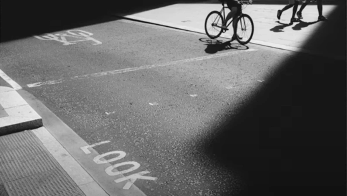 High-contrast black and white photograph of a bicycle and pedestrians on a parking lot with dramatic shadows.