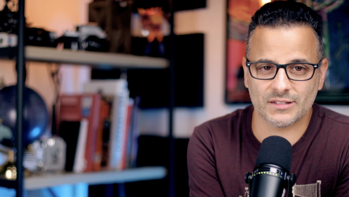 Man wearing glasses and burgundy shirt speaking into a microphone in a home studio setup.