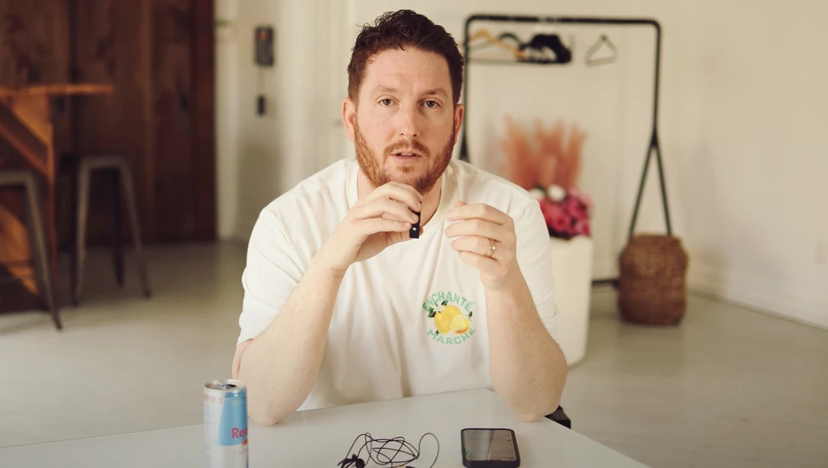 Man in white t-shirt sitting at white table with thoughtful expression, hand near face.