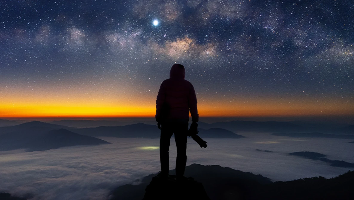 Silhouetted hiker standing on a mountain peak overlooking a sea of clouds at sunrise, with the Milky Way visible in the night sky above.