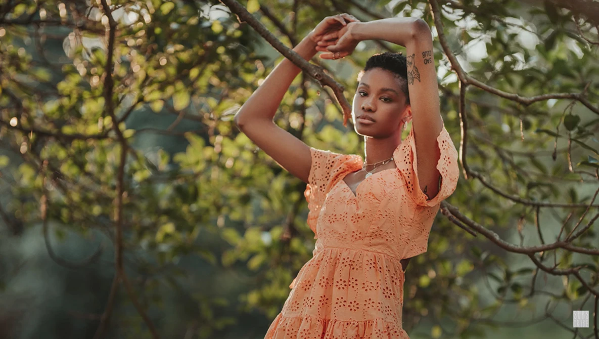 Woman in peach lace dress poses among tree branches with arms raised above head.