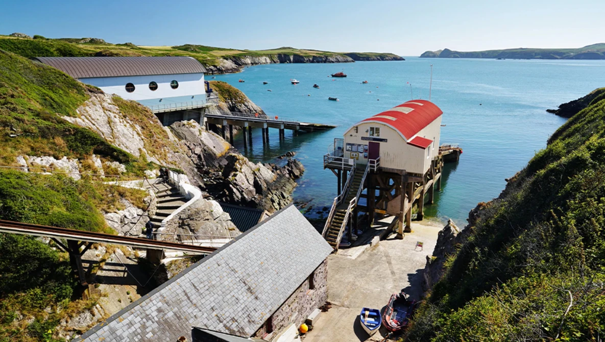 Seaside boathouses with red roofs nestled in a cove surrounded by green cliffs and turquoise water.