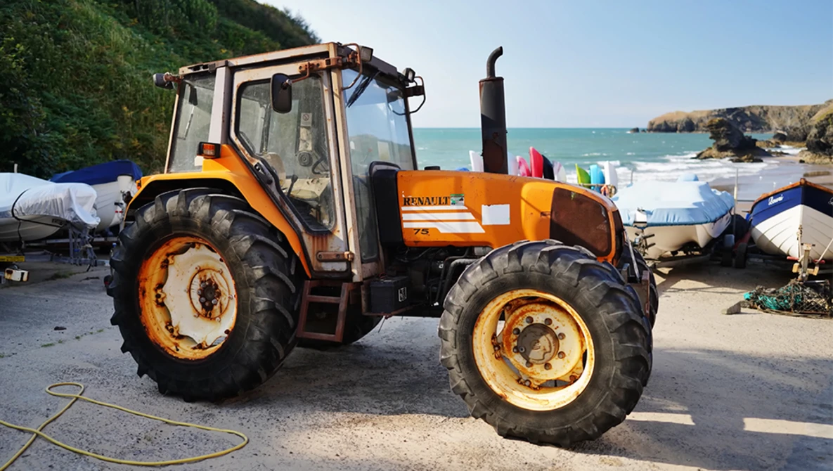 Orange and cream Renault tractor parked at a coastal boat launch, with turquoise water and rocky cliffs visible in the background.
