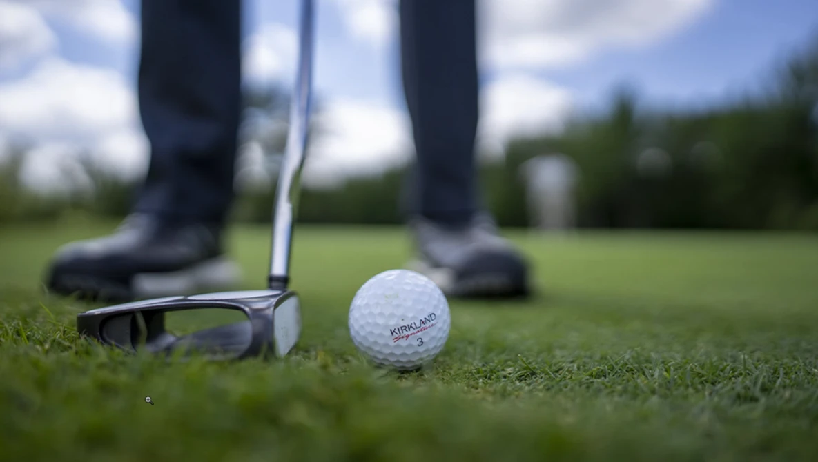 Golf ball on green grass with putter and golfer's feet in background under blue sky.