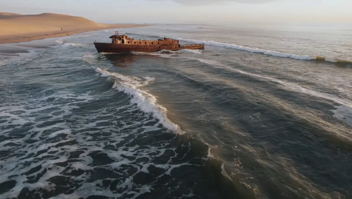 Rusted shipwreck grounded in shallow ocean waters near a sandy desert coastline at sunset.