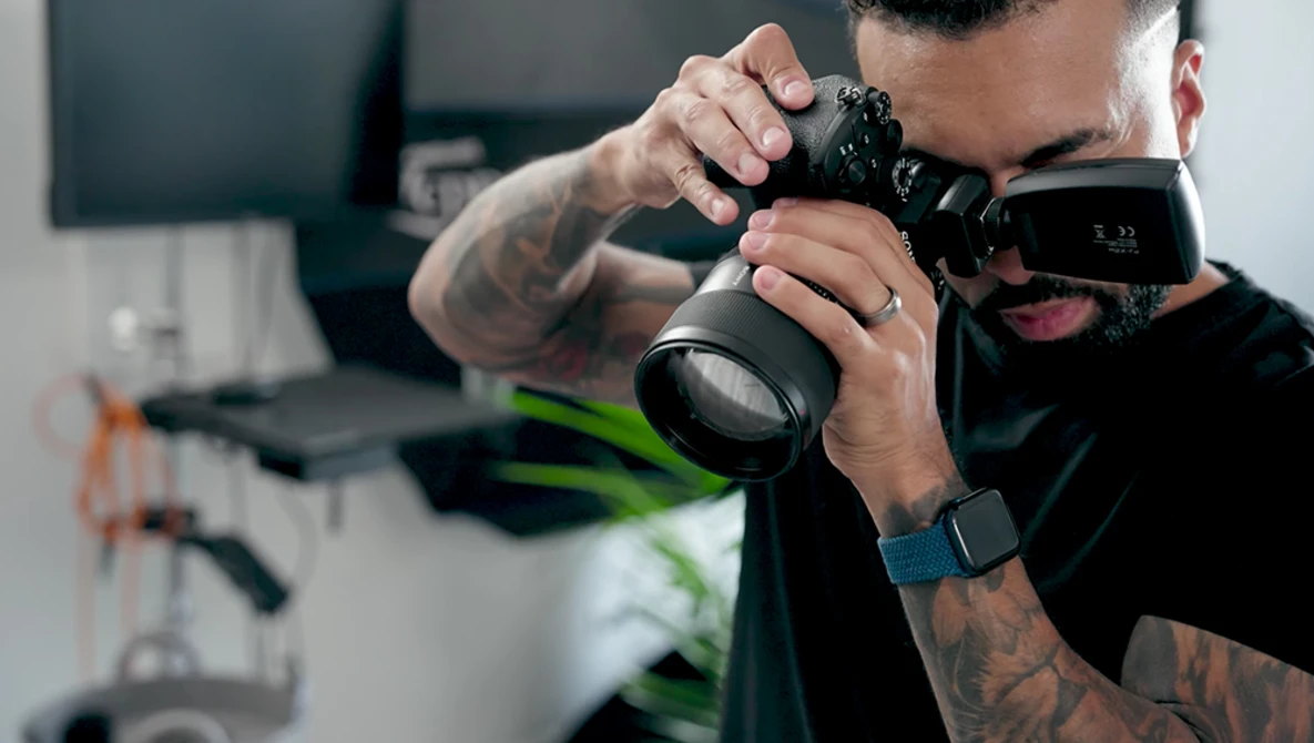 Photographer with tattooed arms holding a mirrorless camera with telephoto lens up to his eye.