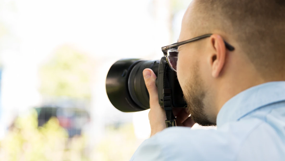 Photographer wearing glasses and light blue shirt holding camera up to eye while shooting outdoors.