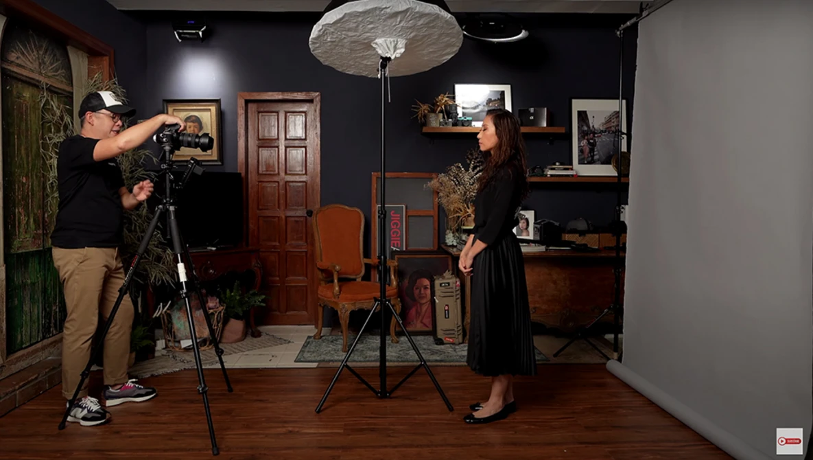 Cameraman on tripod filming a woman in a vintage-styled studio set with warm wood tones and warm lighting.