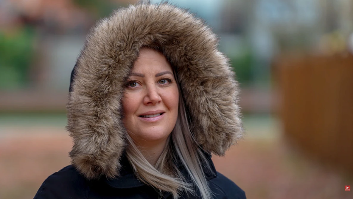 Woman wearing a black jacket with a large fur-lined hood, photographed outdoors with a soft-focus background.