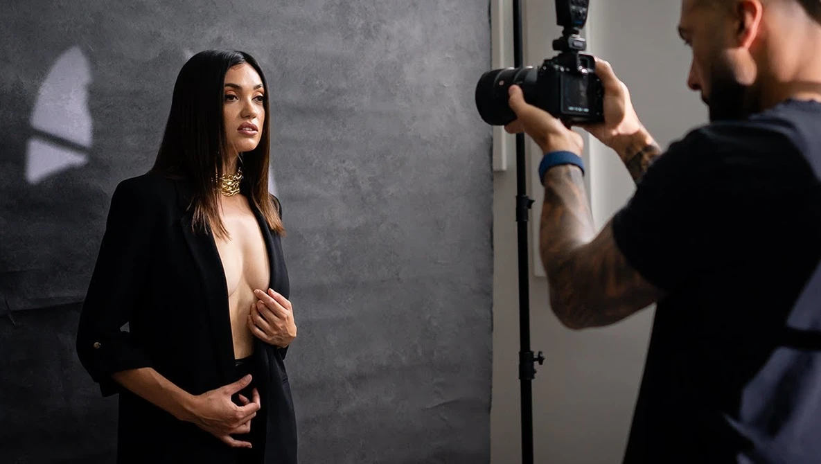 Photographer holding camera on tripod while female subject poses against gray backdrop during studio portrait session.