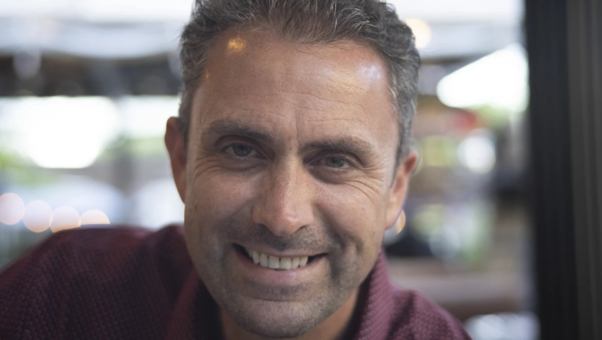 Middle-aged man with gray hair smiling at camera in indoor setting with blurred background.