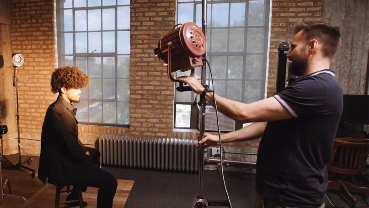 Photographer adjusting a studio light on a tripod while a woman sits for a portrait in an industrial loft space.