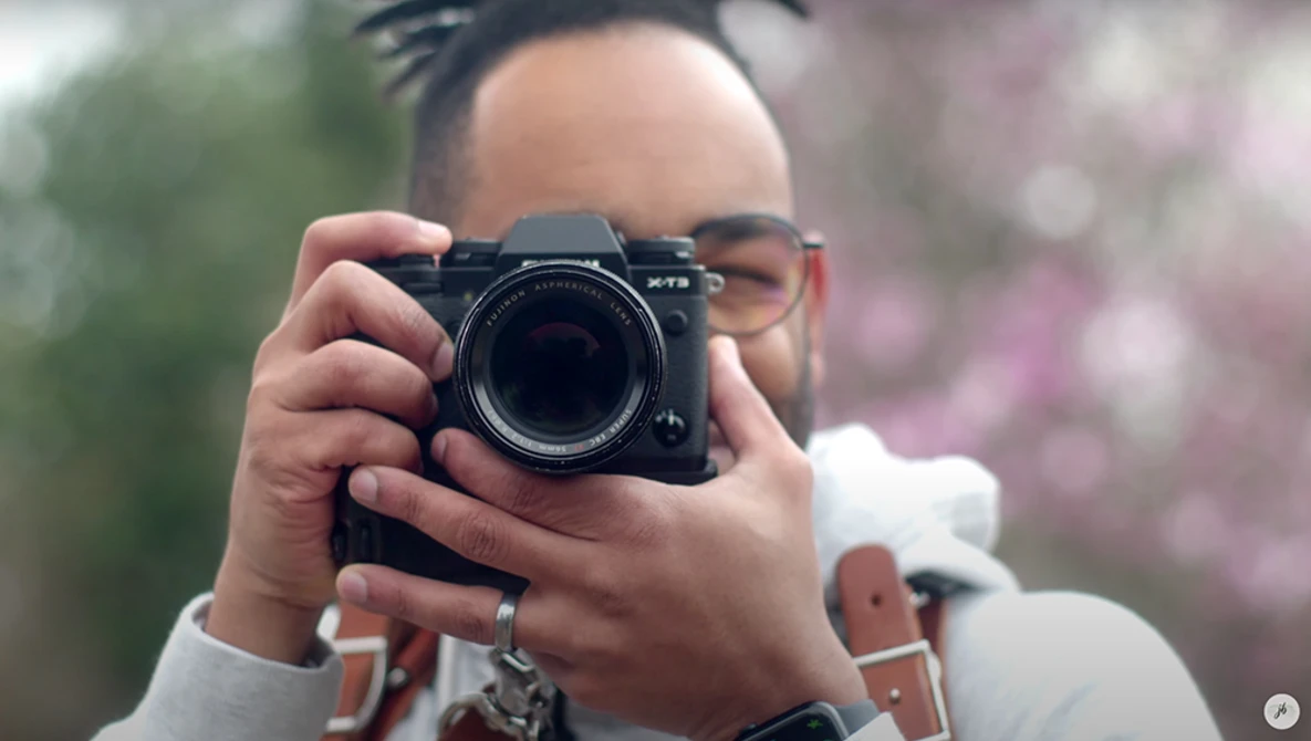 Photographer holding a mirrorless camera up to take a photo, wearing glasses and a white long-sleeve shirt.