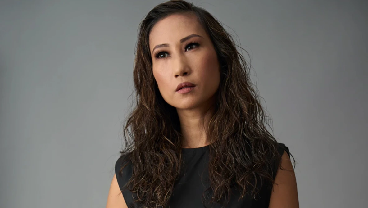 Woman with long dark wavy hair wearing sleeveless black top against neutral gray background.