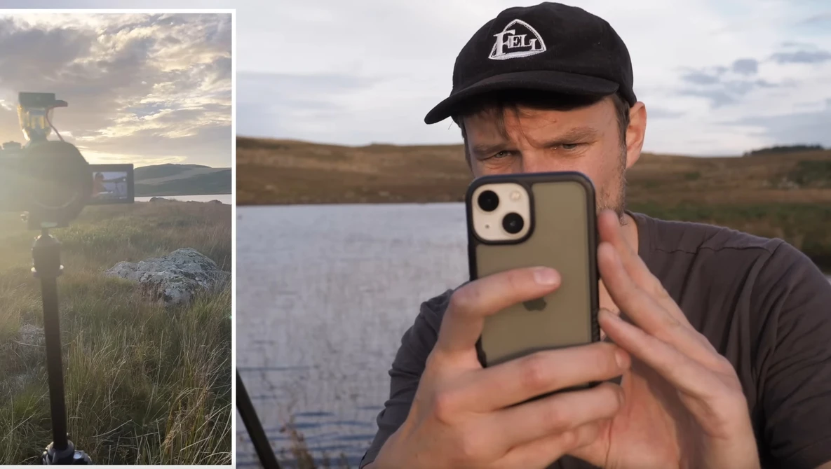 Photographer holding smartphone taking selfie outdoors beside water during golden hour.