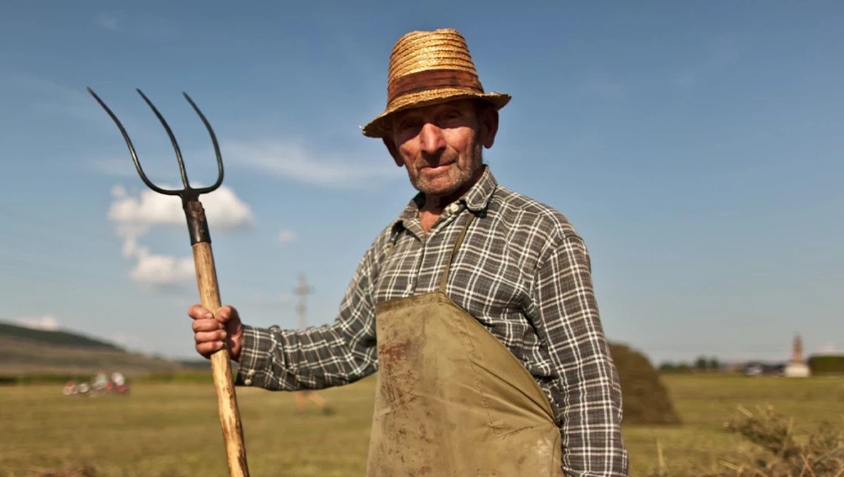 Farmer in straw hat and work apron holding a pitchfork in an open field.