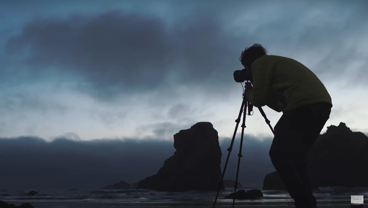 Photographer silhouetted against dramatic cloudy sky while operating tripod-mounted camera on rocky coastal terrain.