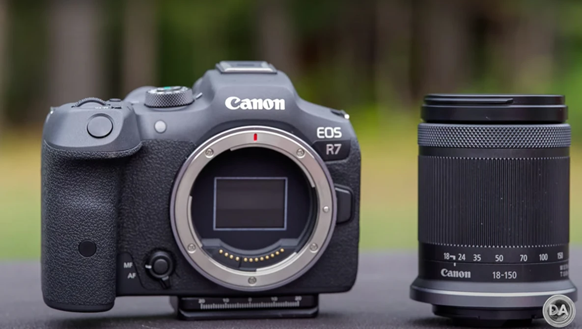Canon EOS R7 mirrorless camera body with Canon 18-150mm lens displayed against blurred outdoor background.