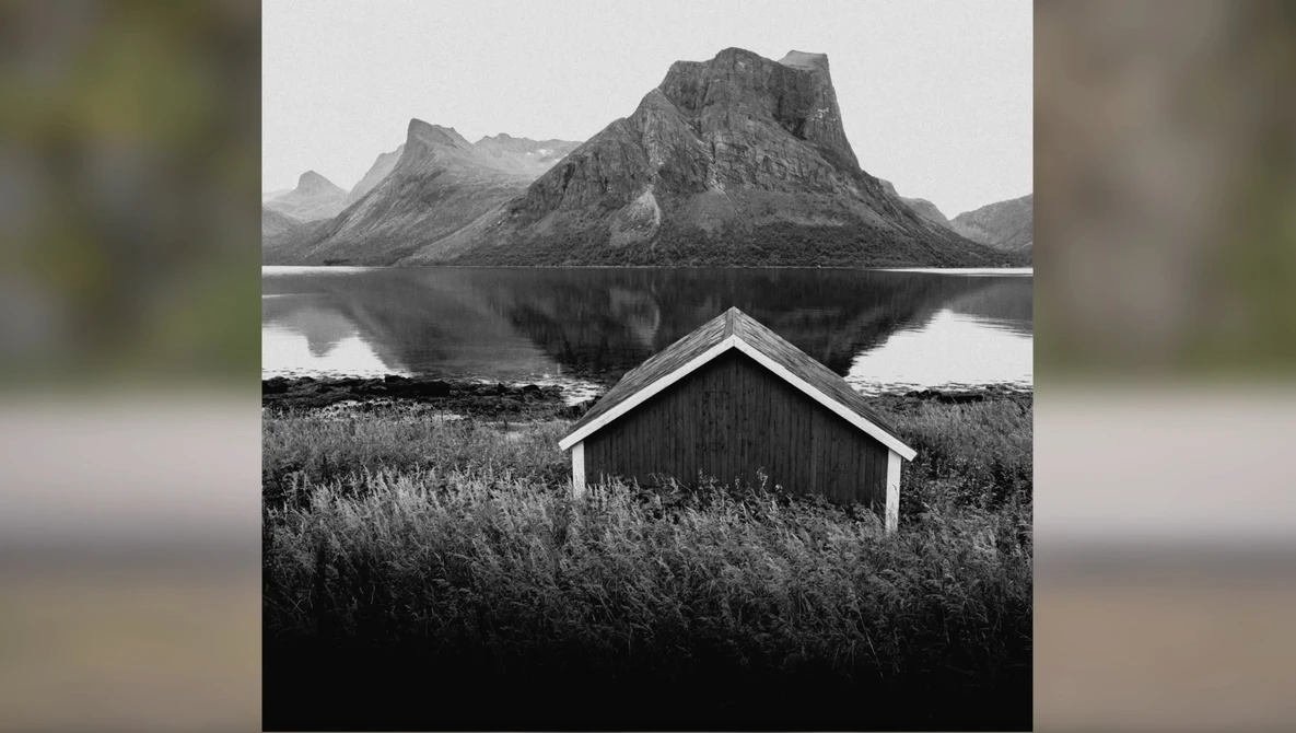 Minimalist black and white landscape with a small wooden boathouse reflected in still water, mountains rising in the background.